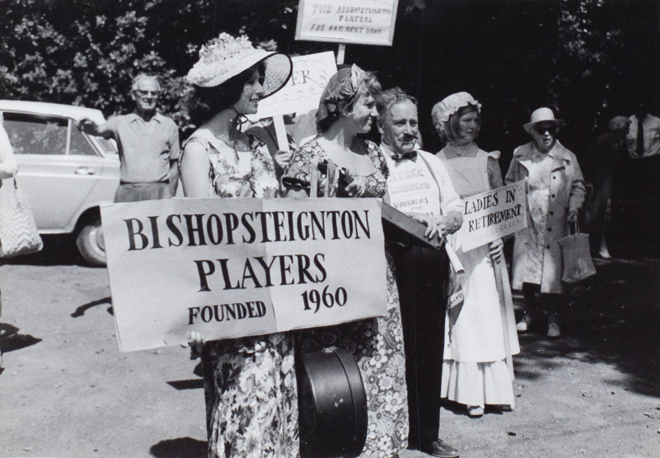 Photograph of some of the Bishopsteignton Players at the Church Fete 1974 showing a banner 'Bishopsteignton Players Founded 1960' and a poster 'Ladies in Retirement'.